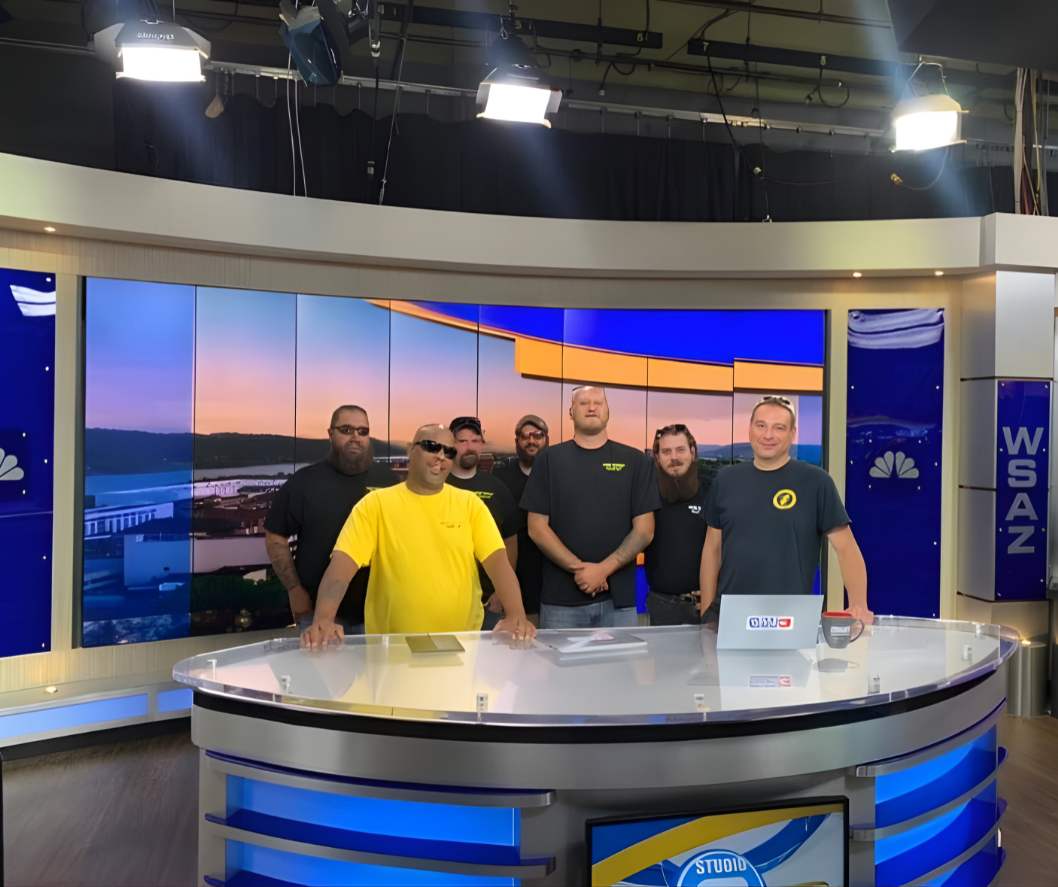 A group of six men standing behind a news studio desk with a large screen displaying a scenic landscape with a river and sunset in the background.