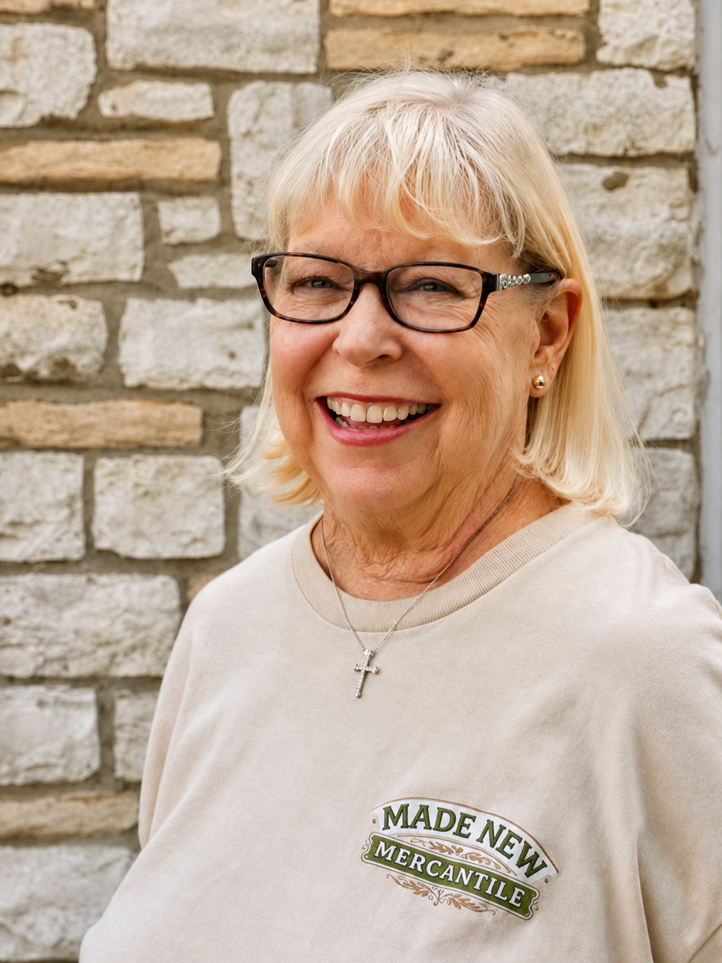 Smiling older woman with blonde hair, glasses, and a cross necklace posing in front of a brick wall, wearing a beige shirt with 'Made New Mercantile' logo.