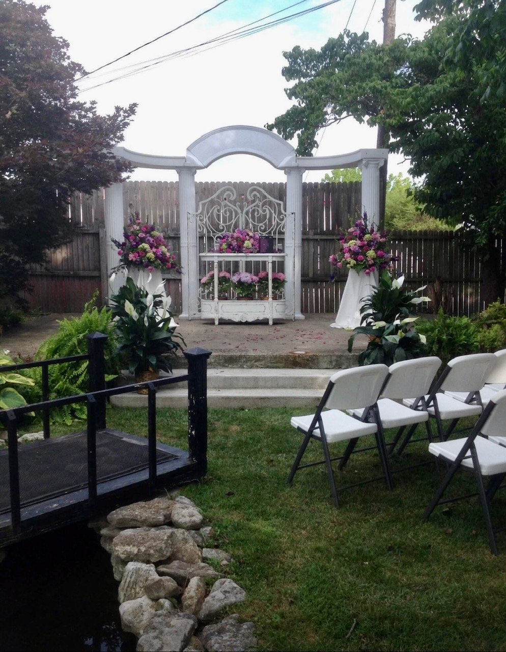 Outdoor wedding altar with white wrought iron gate, floral arrangements in pink, purple, and white, on a small stage with white chairs arranged for guests, wooden fence background, small pond with rocks, and lush green trees.
