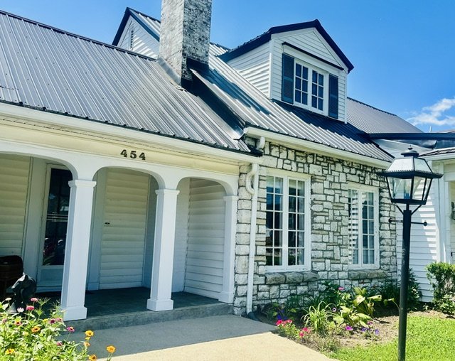 Front view of a house with a stone and wood exterior, metal roof, dormer window, arched porch, house number 454, and a lamp post near the sidewalk, with flower beds and greenery.