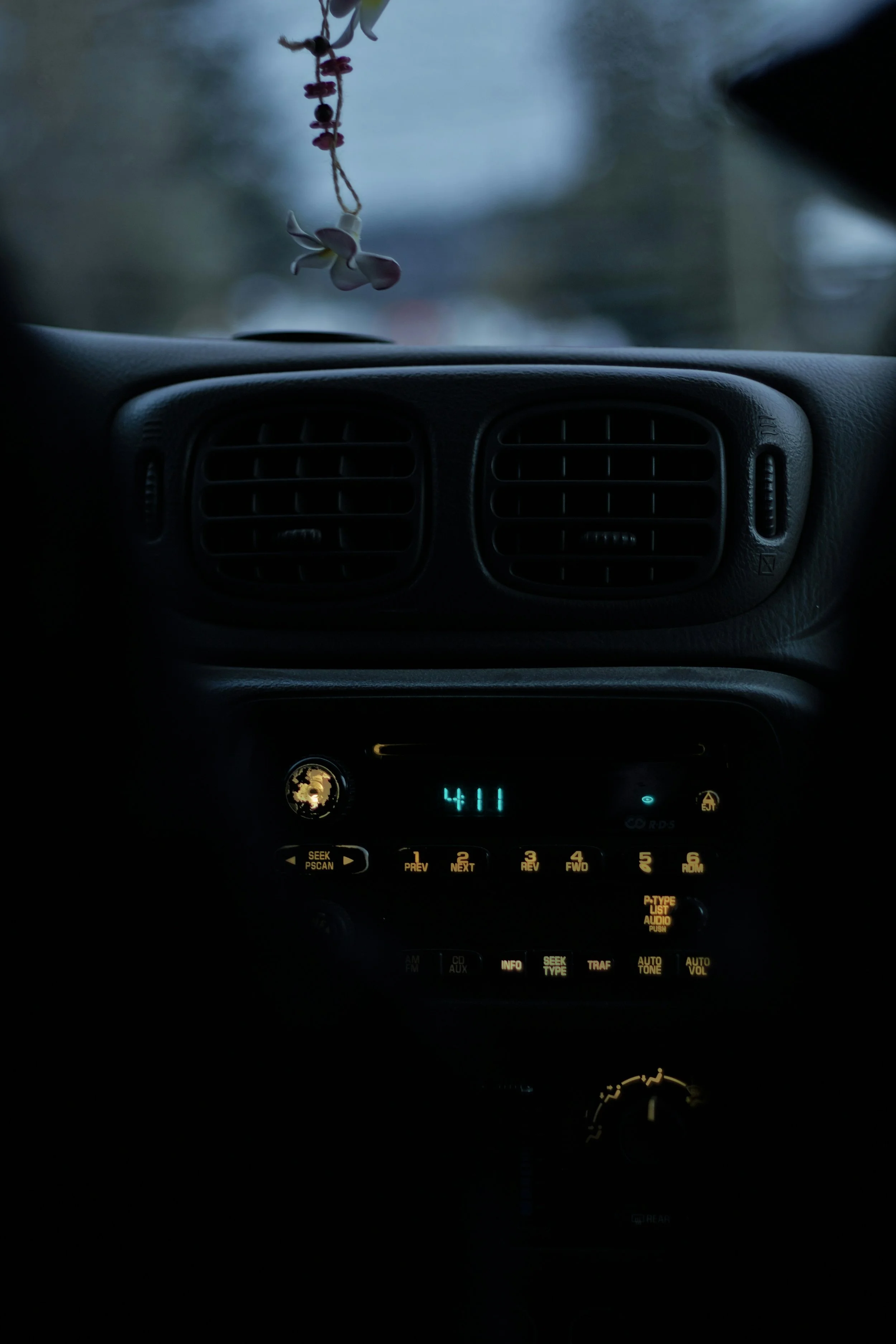 The image shows a car dashboard view through the windshield. It features a digital clock displaying 4:11, a radio panel with yellow-lit buttons and labels, and two front air vents. The dashboard appears dark with a slight glow from the digital and button lights. The outside scene is blurry with a cloudy sky, and there are some decorations hanging from the rearview mirror.