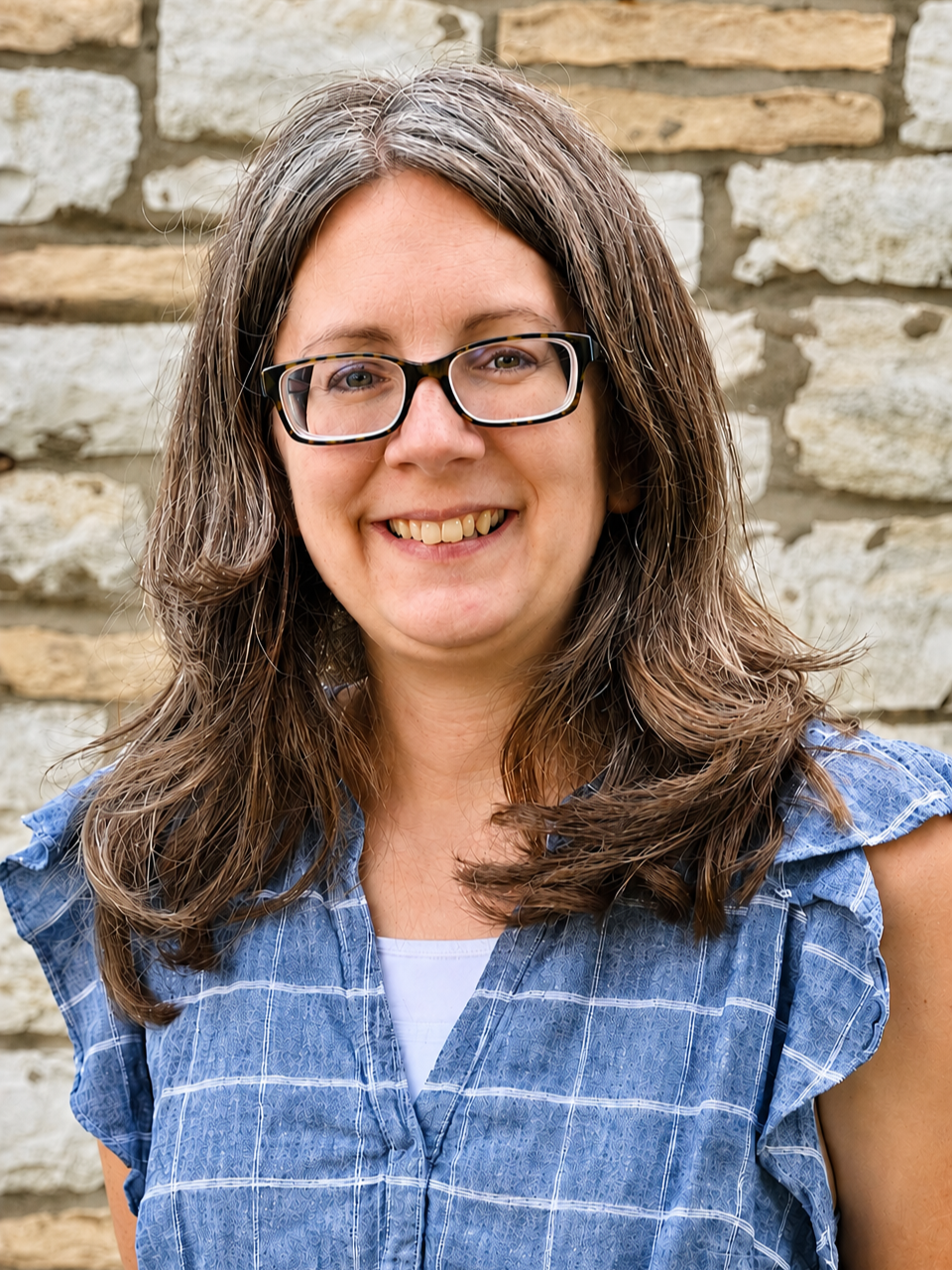 A woman with long brown hair, wearing glasses and a blue plaid shirt, smiling in front of a stone wall.