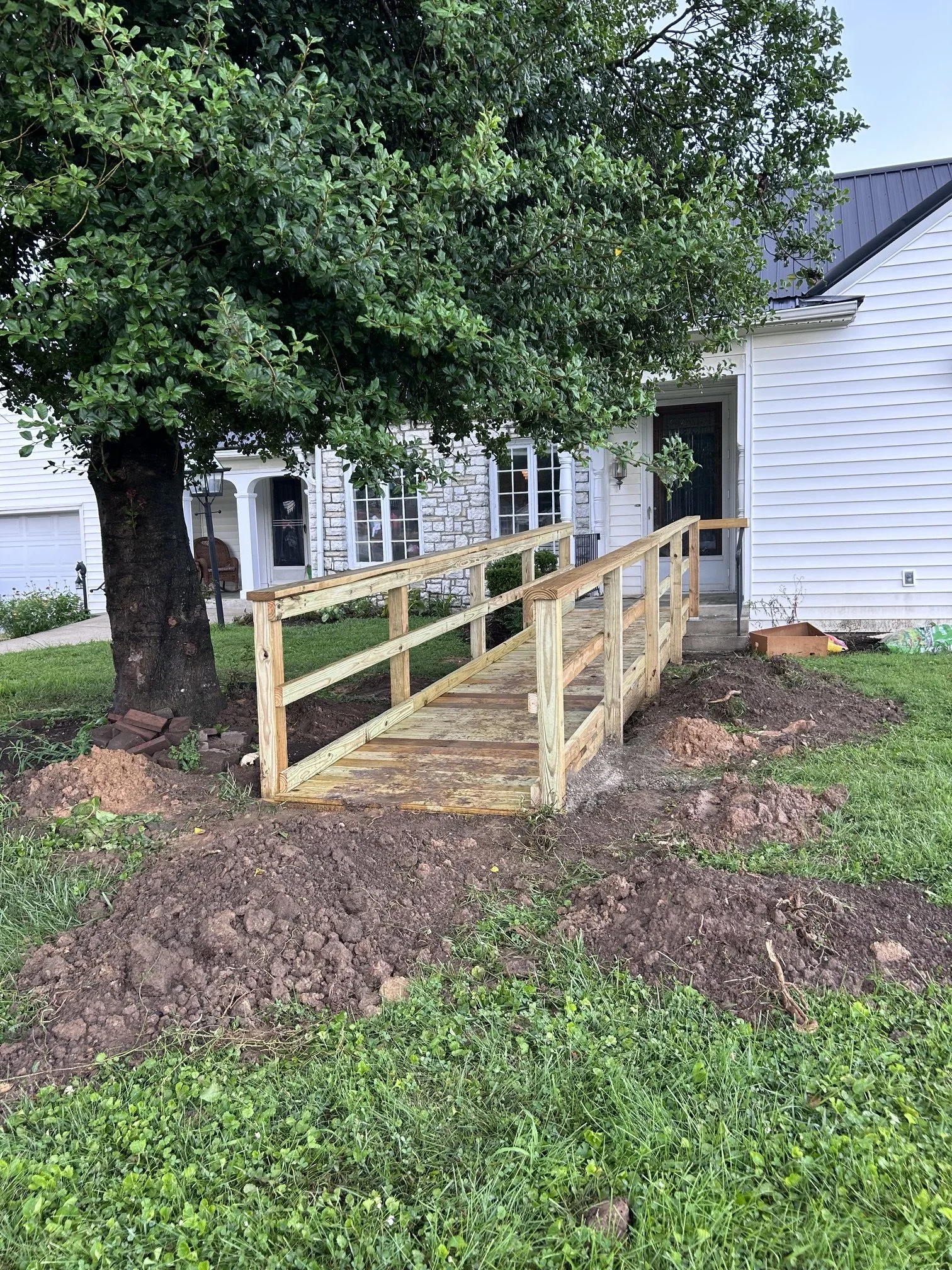 A wooden ramp leads to the front door of a house, which has white siding and a stone facade near the entrance. A large leafy tree shades part of the yard. The ground around the ramp is dug up, with dirt and patches of grass visible.