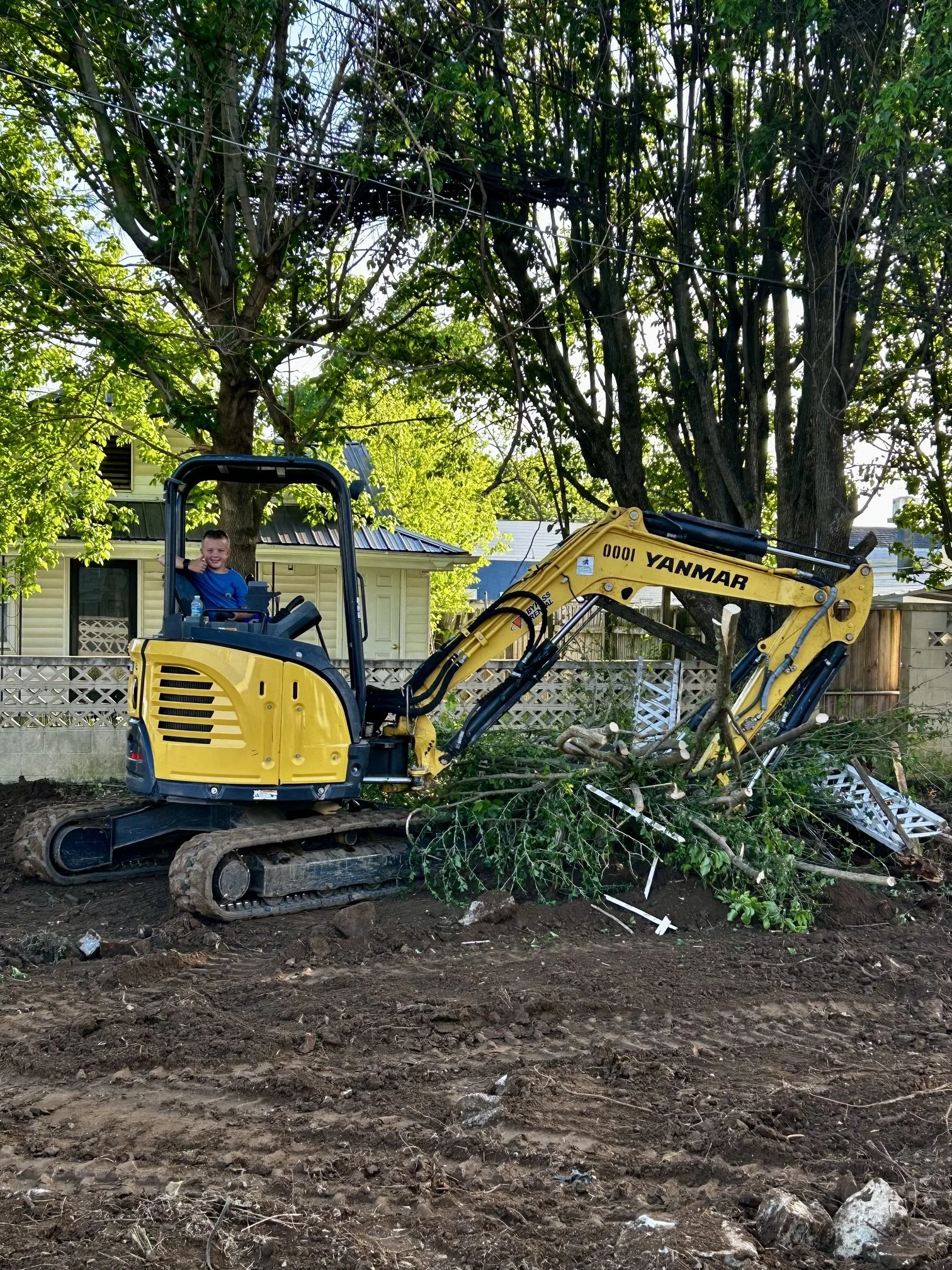 A young boy sitting in a yellow excavator, smiling and giving a thumbs-up, with freshly dug earth and debris in the foreground, a house in the background, and several large trees with green leaves and branches.