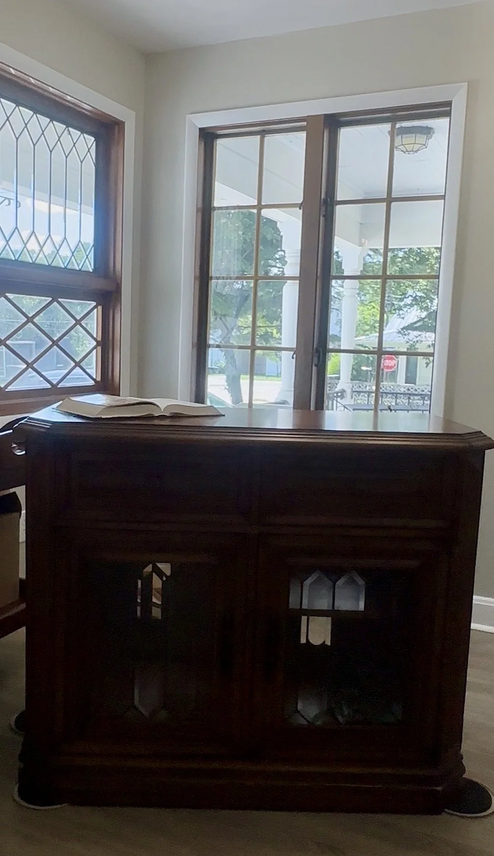 A wooden cabinet with a book on top, located in front of two windows with wooden frames, one with diamond-patterned leaded glass and the other with clear glass panes. Outside, a porch and trees are visible.