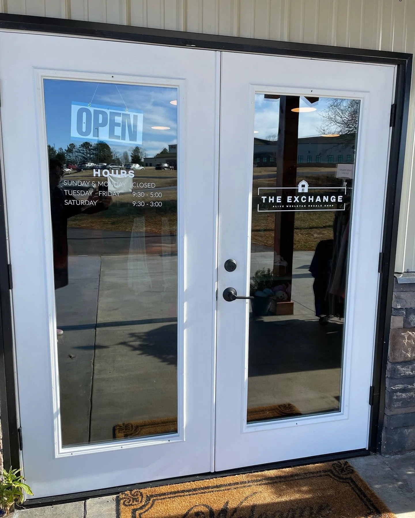 Double glass door with an 'Open' sign at the top left, business hours listed below, and a logo with the name 'The Exchange' on the right door, reflecting the outside parking lot and trees.