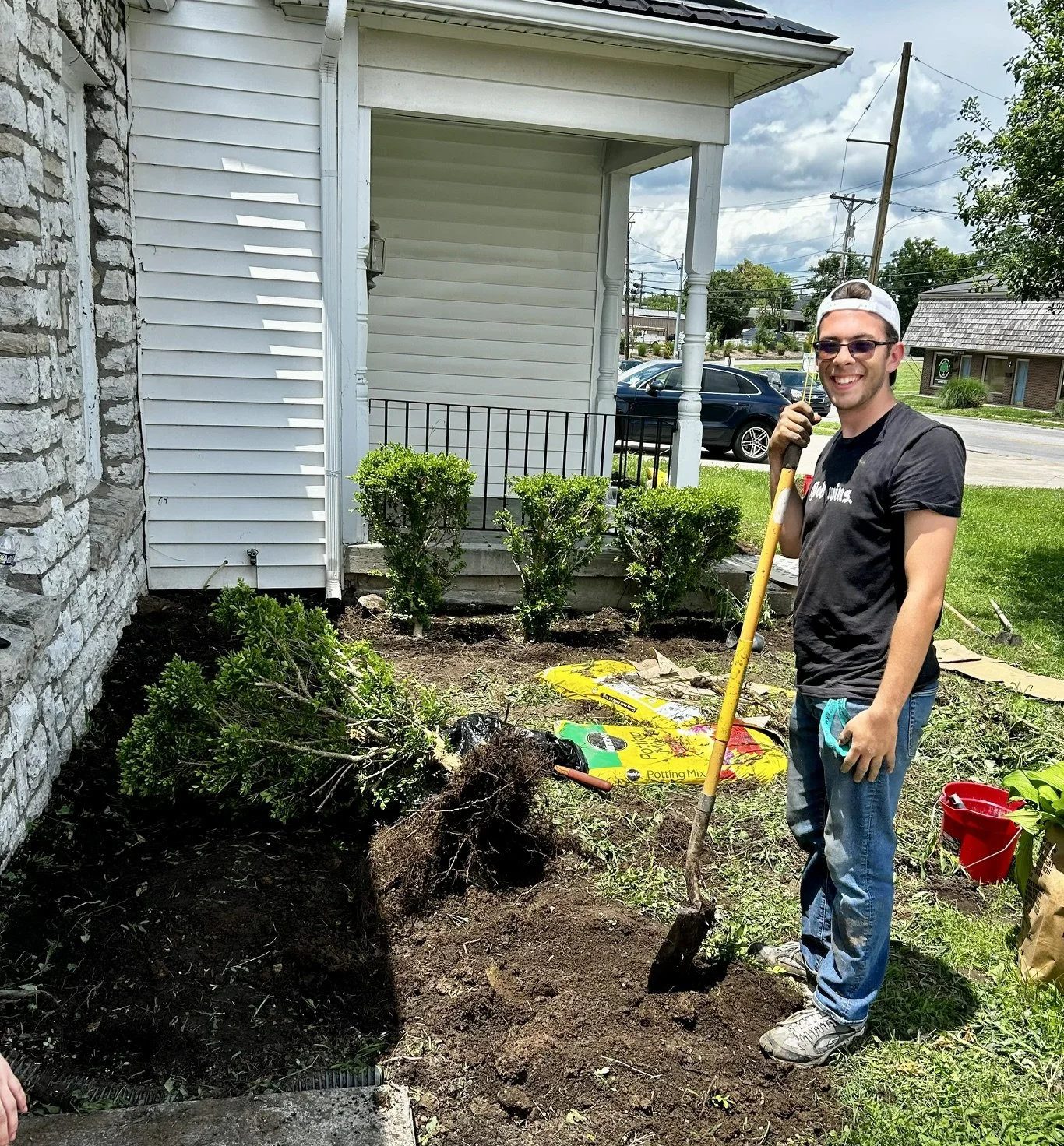 A young man smiling outdoors holds a shovel while gardening near a house. There are shrubs, garden soil, a bag of potting mix, and gardening tools around him.