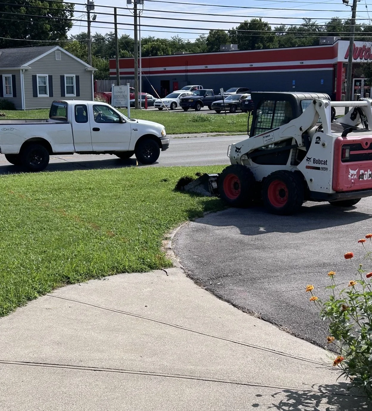 A white pickup truck parked on the side of a street next to a small Bobcat skid-steer loader on a paved driveway, with a grassy yard and sidewalk in the foreground. In the background, there is a commercial building, several parked cars, and utility poles with power lines.