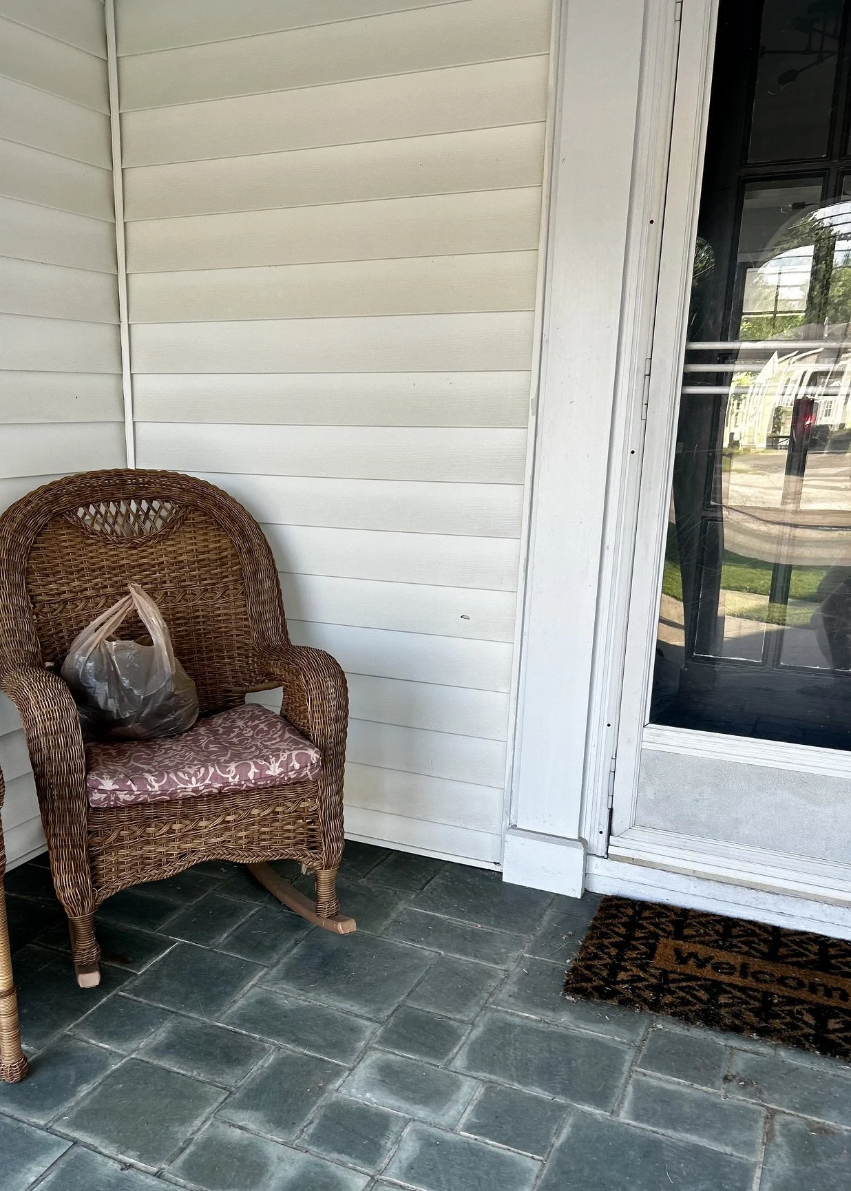 A wicker armchair with a pink cushion, a plastic bag on it, on a porch with gray tile flooring, white siding, and a glass door with a white frame.
