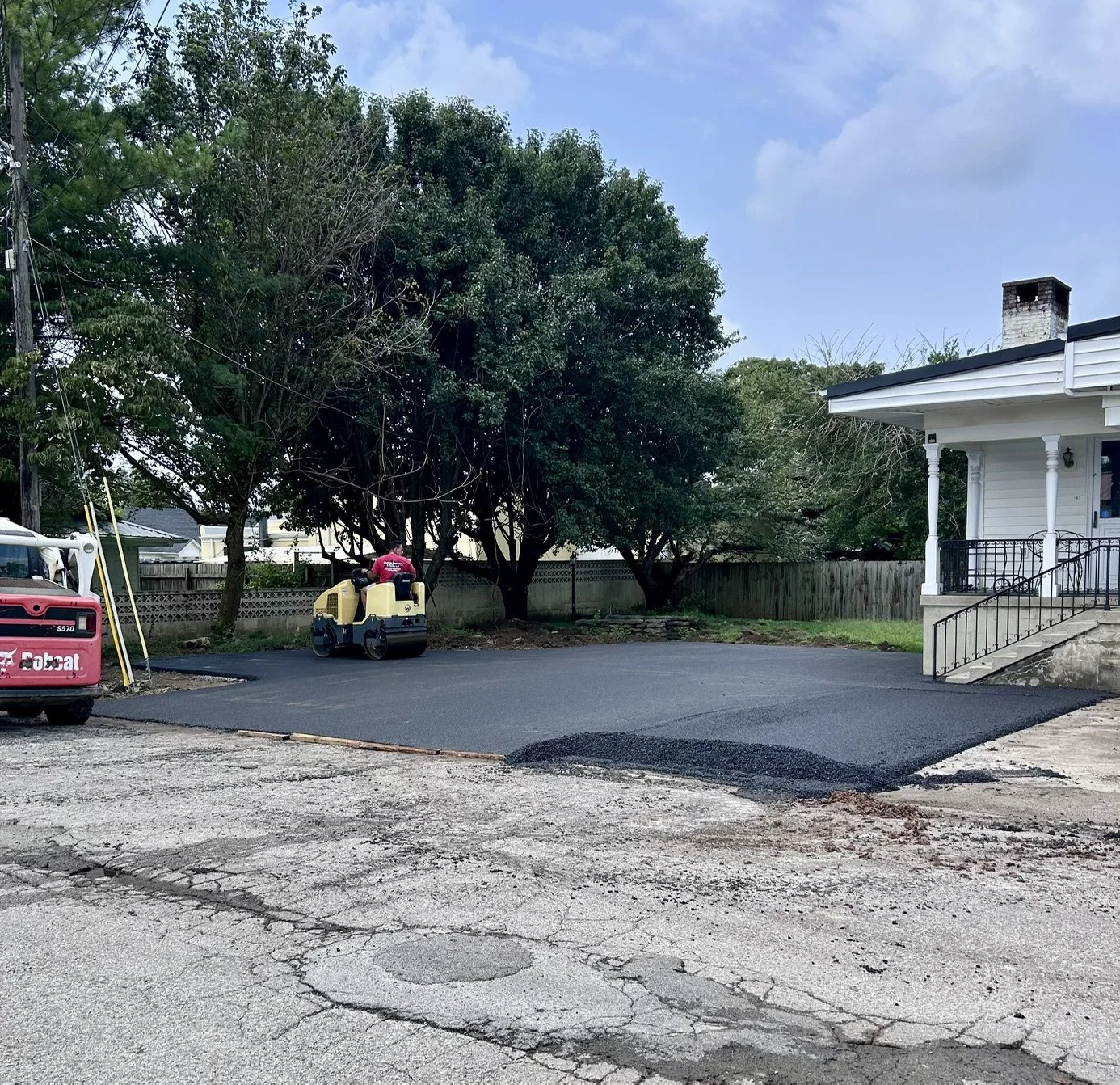Residential backyard with fresh asphalt being laid, a roller compactor, and construction equipment, large trees, a wooden fence, a house with porch and stairs, and a partly cloudy sky.