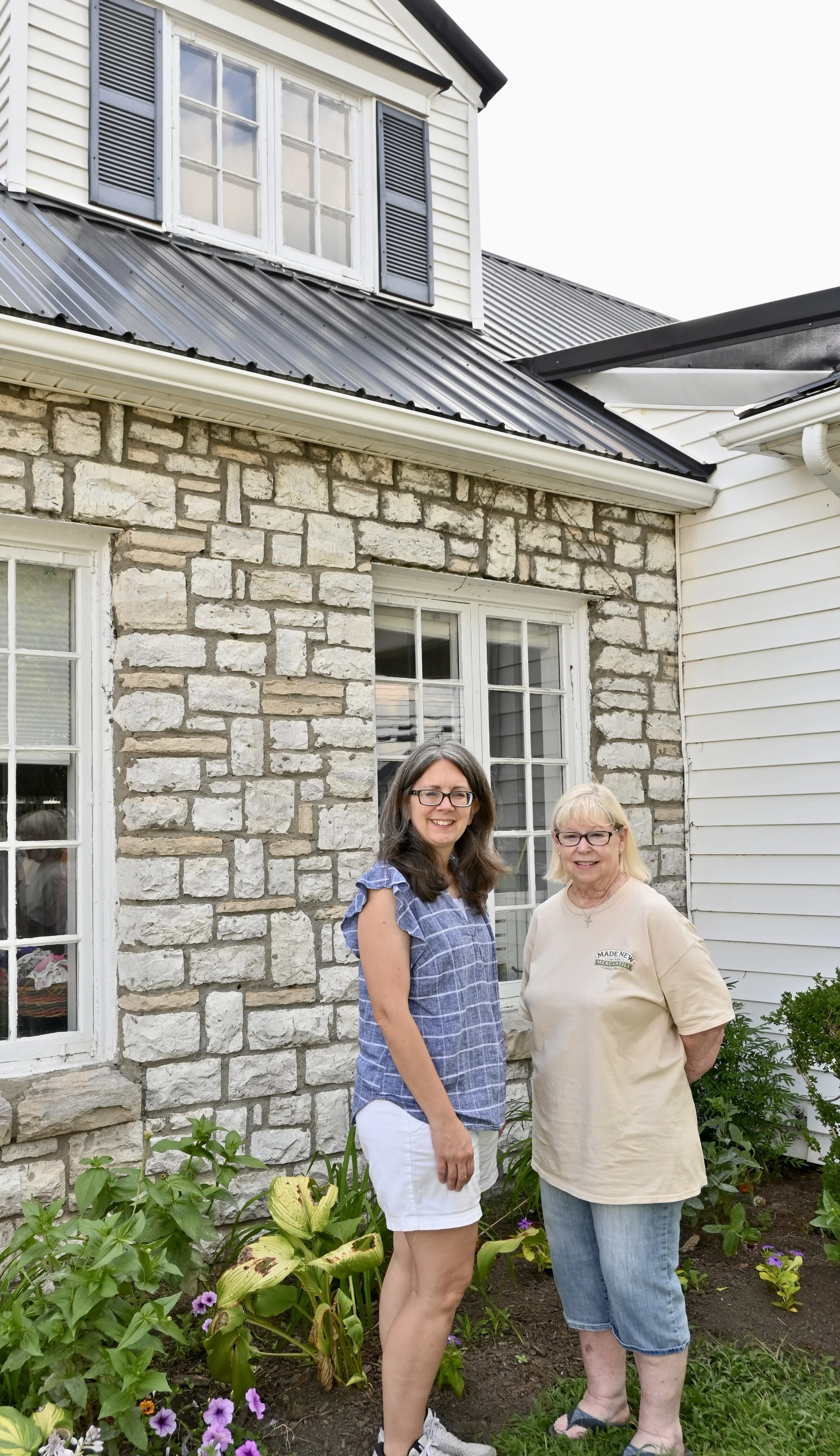 Two women standing in front of a house with stone and white siding, in a garden with green plants and purple flowers.