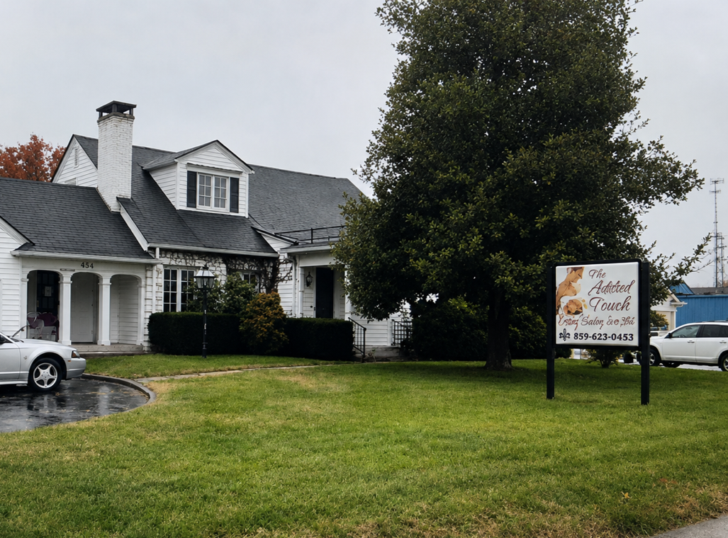 A white, two-story house with black roof, chimney, and front porch, surrounded by a lawn and trees, with a sign for The Addicted Touch Spray Salon outside.