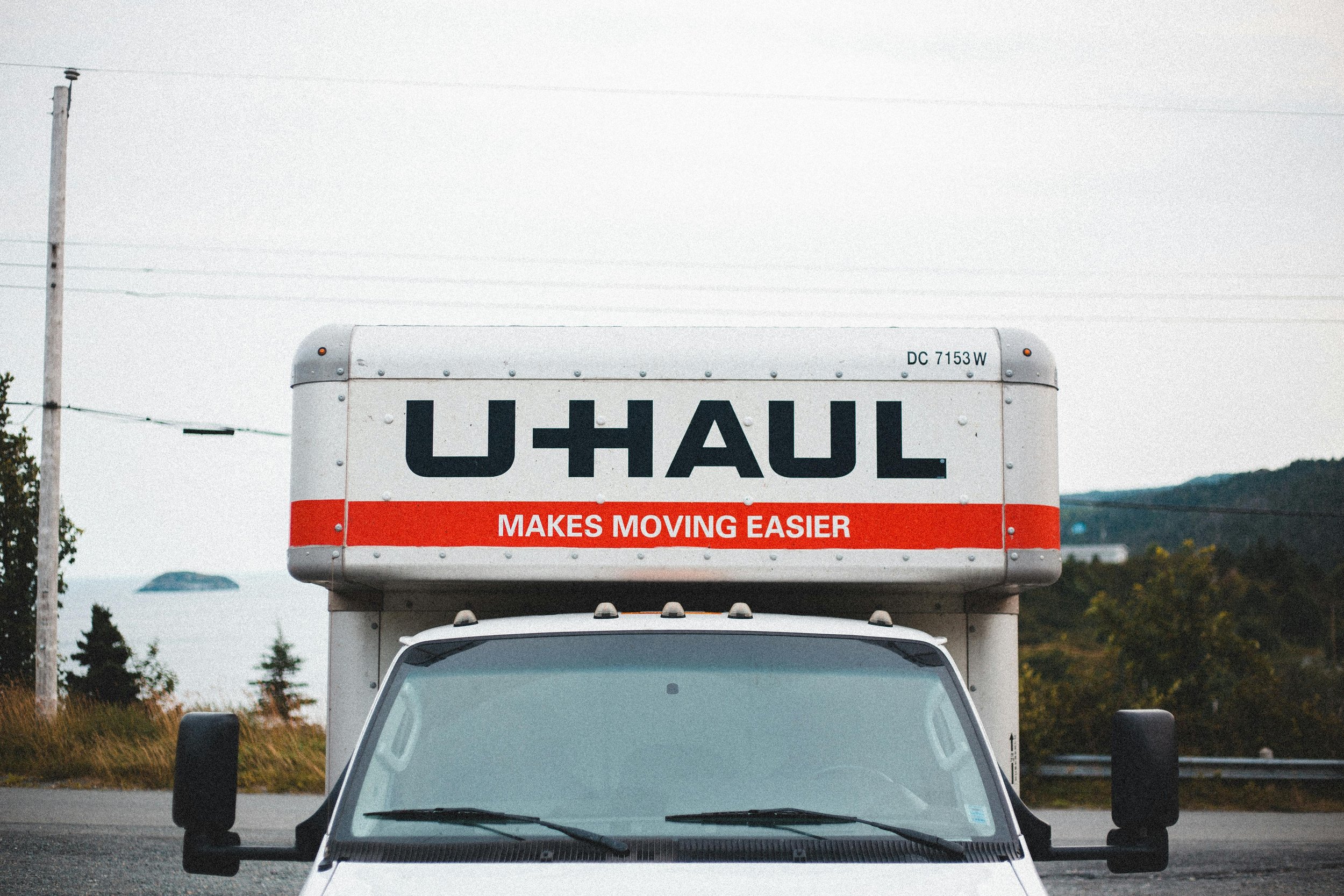 The front of a U-Haul moving truck with part of the cab visible, parked outdoors with trees and a hill in the background.