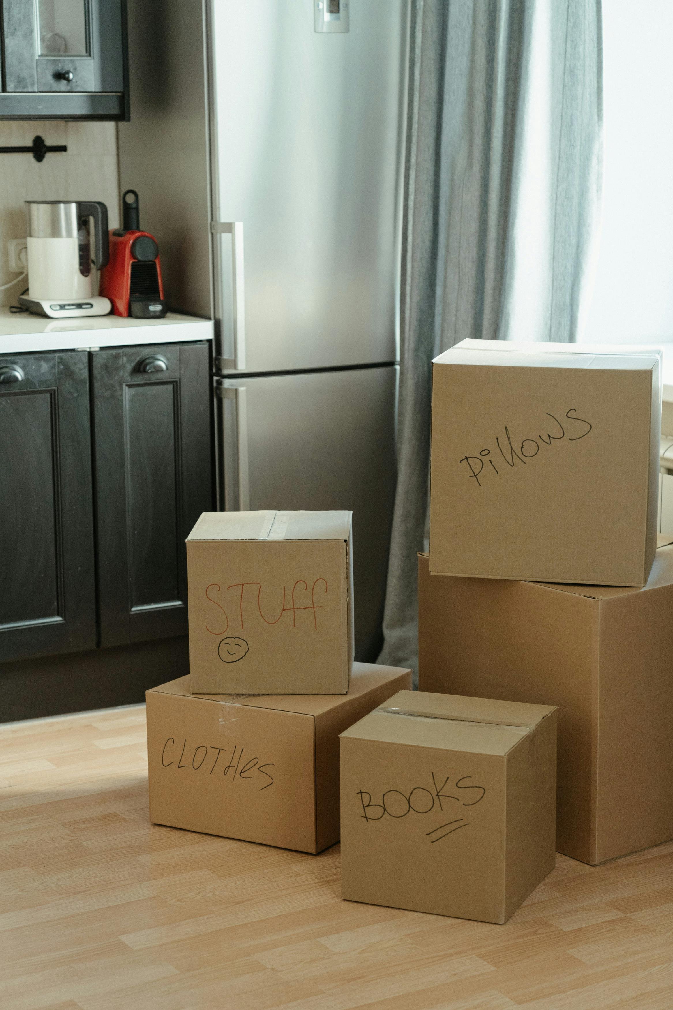 Several cardboard boxes labeled as 'STUFF,' 'CLOTHES,' 'BOOKS,' and 'PILLOWS,' stacked in a room with a kitchen area in the background, including a refrigerator, coffee maker, and curtains.