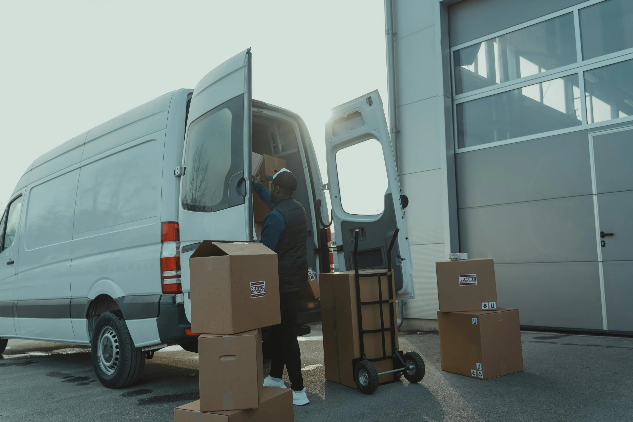 A person loading cardboard boxes into a white delivery van outside a building with large windows. The boxes are labeled fragile, and some are on a dolly. Going to Nashville