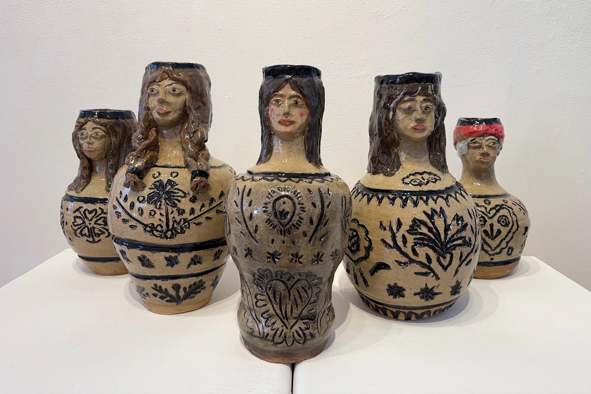 Five ceramic vases with faces and hand-painted black designs on a white surface against a plain wall.