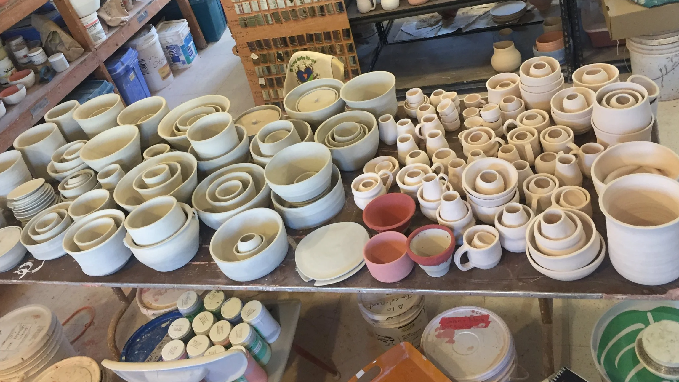 A large table filled with various unglazed ceramic bowls, cups, and plates in different sizes, some pink-colored, in a pottery studio setting.