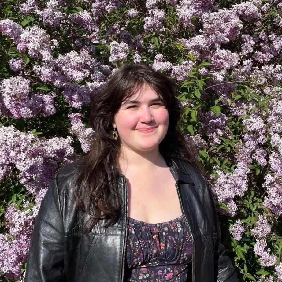 A young woman with dark brown hair smiling outdoors, standing in front of a large bush filled with pink and purple flowers.