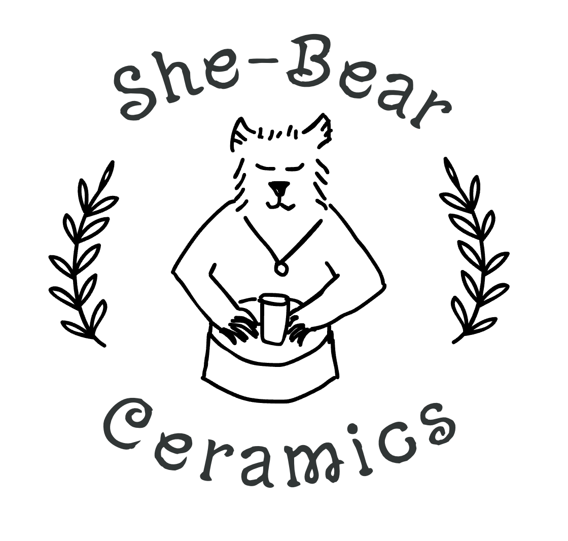 A logo featuring a bear with closed eyes, wearing an apron and holding a cup, surrounded by two leafy branches. The text 'she-Bear ceramics' is written above and below the bear.