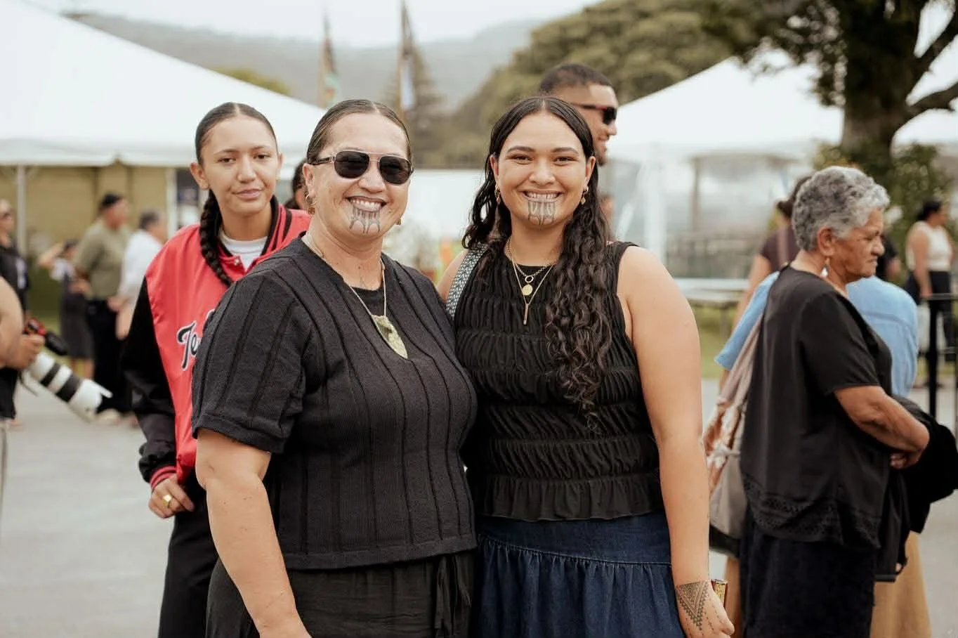 Three women smiling at an outdoor event, with tents and other people in the background.
