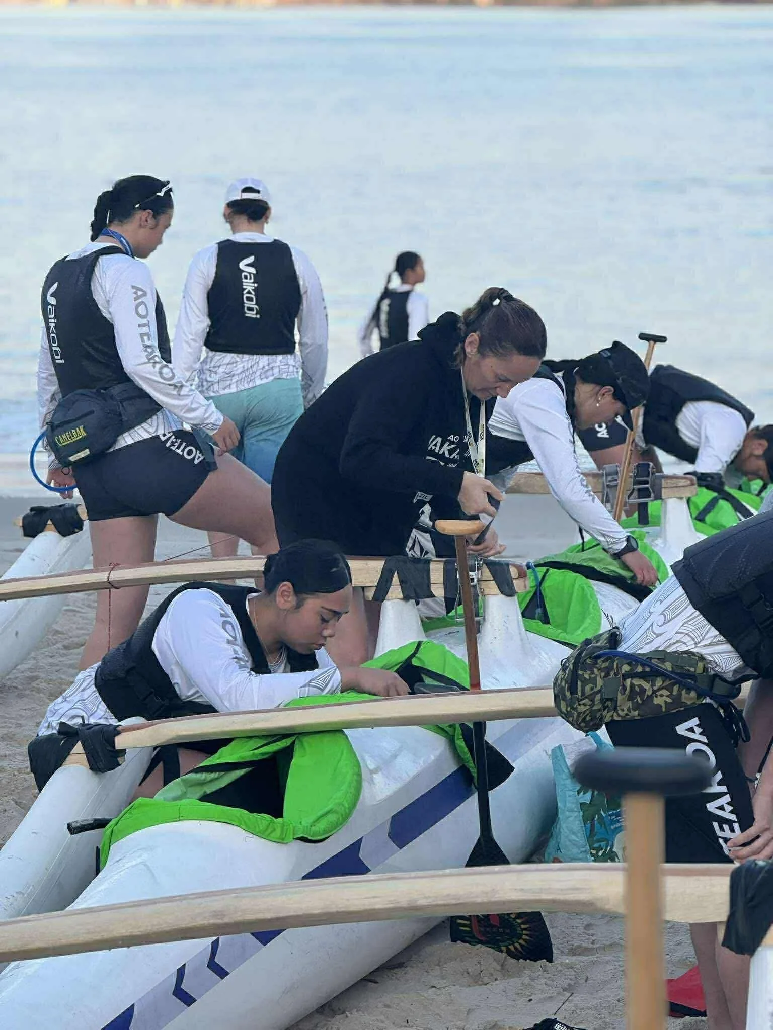Group of women preparing dragon boats on a beach for racing, some are adjusting equipment while others stand nearby.