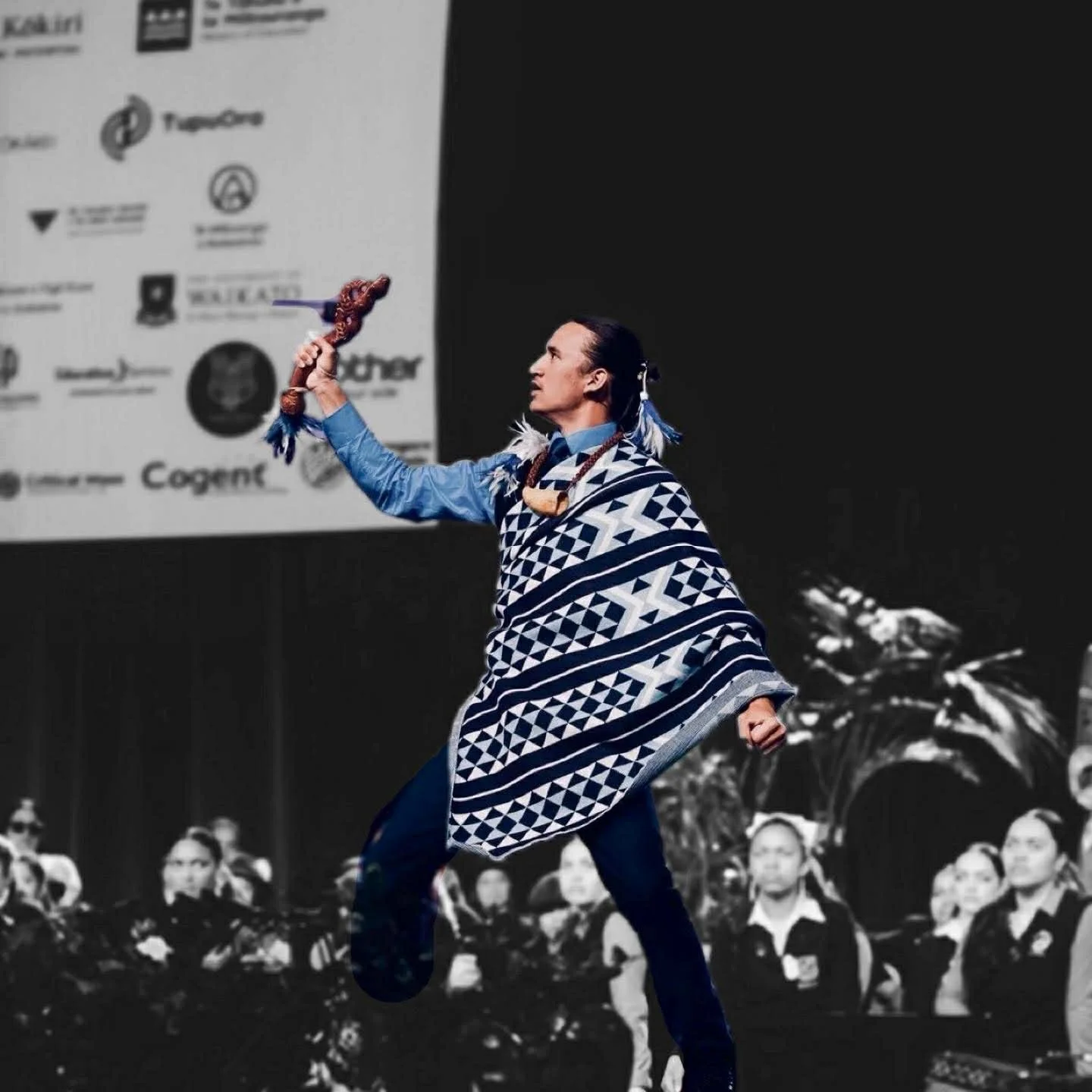 A man dressed in traditional indigenous Maasai attire holding a ceremonial spear, performing on stage with an audience and a large screen in the background.