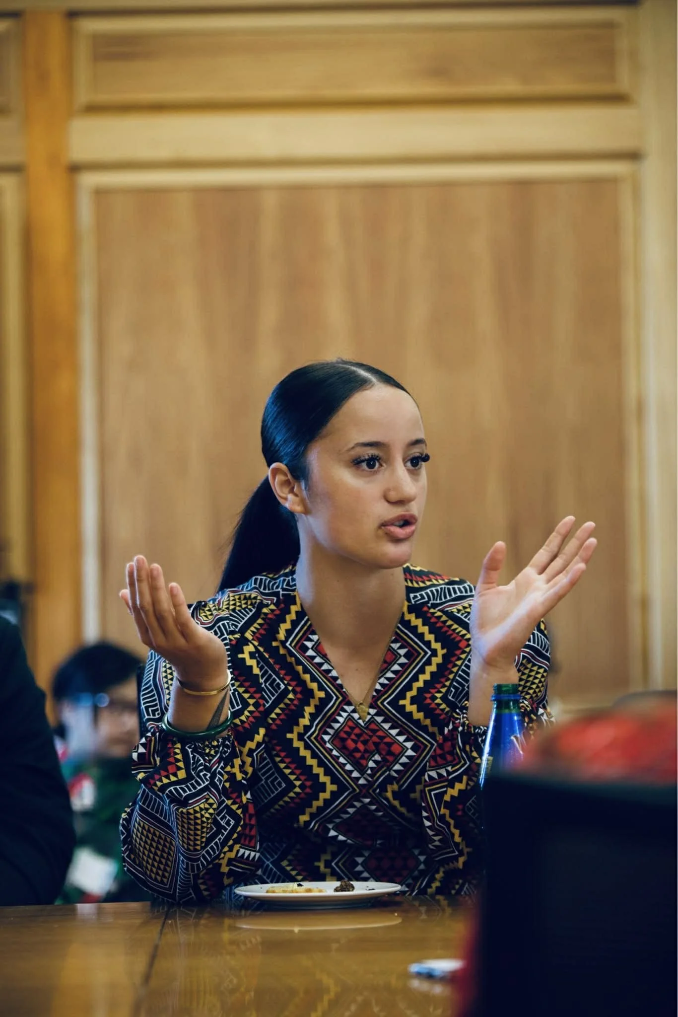 A young woman with dark hair in a ponytail, wearing a colorful patterned shirt, is sitting at a table with a plate and a water bottle in front of her, gesturing with her hands as she talks.
