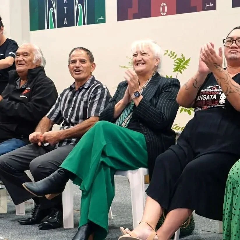 Four adults seated and clapping during an indoor event, with a colorful artwork on the wall behind them.