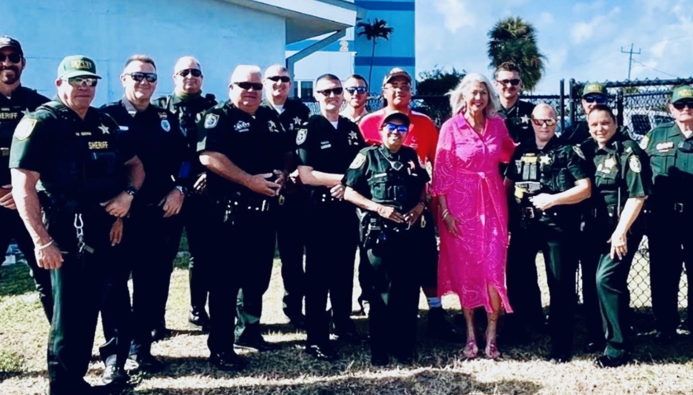 Group of police officers and civilians standing outdoors on a sunny day, with a blue building, palm trees, and power lines in the background.