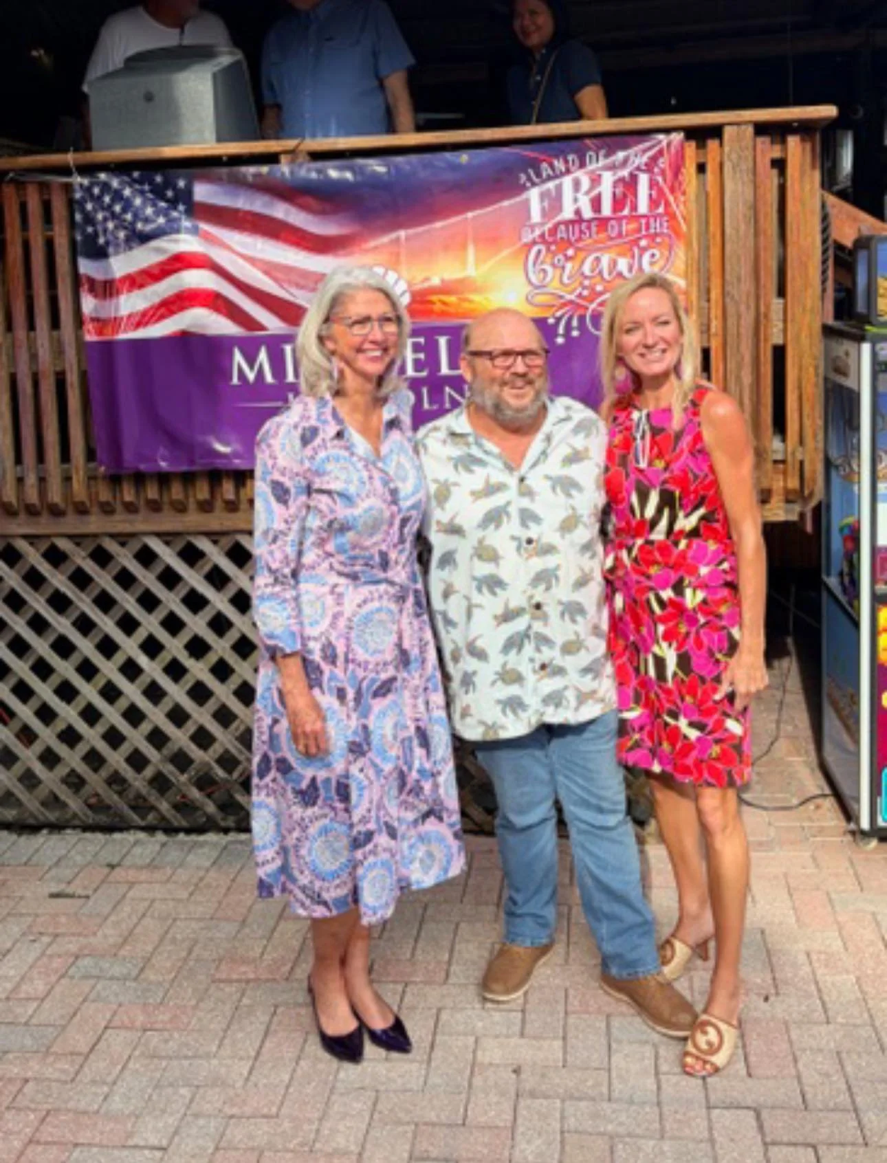 Three people standing outdoors in front of a colorful banner with American flag and sunset design. The banner reads "Land of the Free Because of the Brave." The group includes two women and one man, all smiling, dressed in casual, summery clothing.