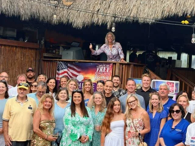 Group of people posing outdoors with a woman standing behind a bar or balcony, giving a thumbs-up, at a festive event.