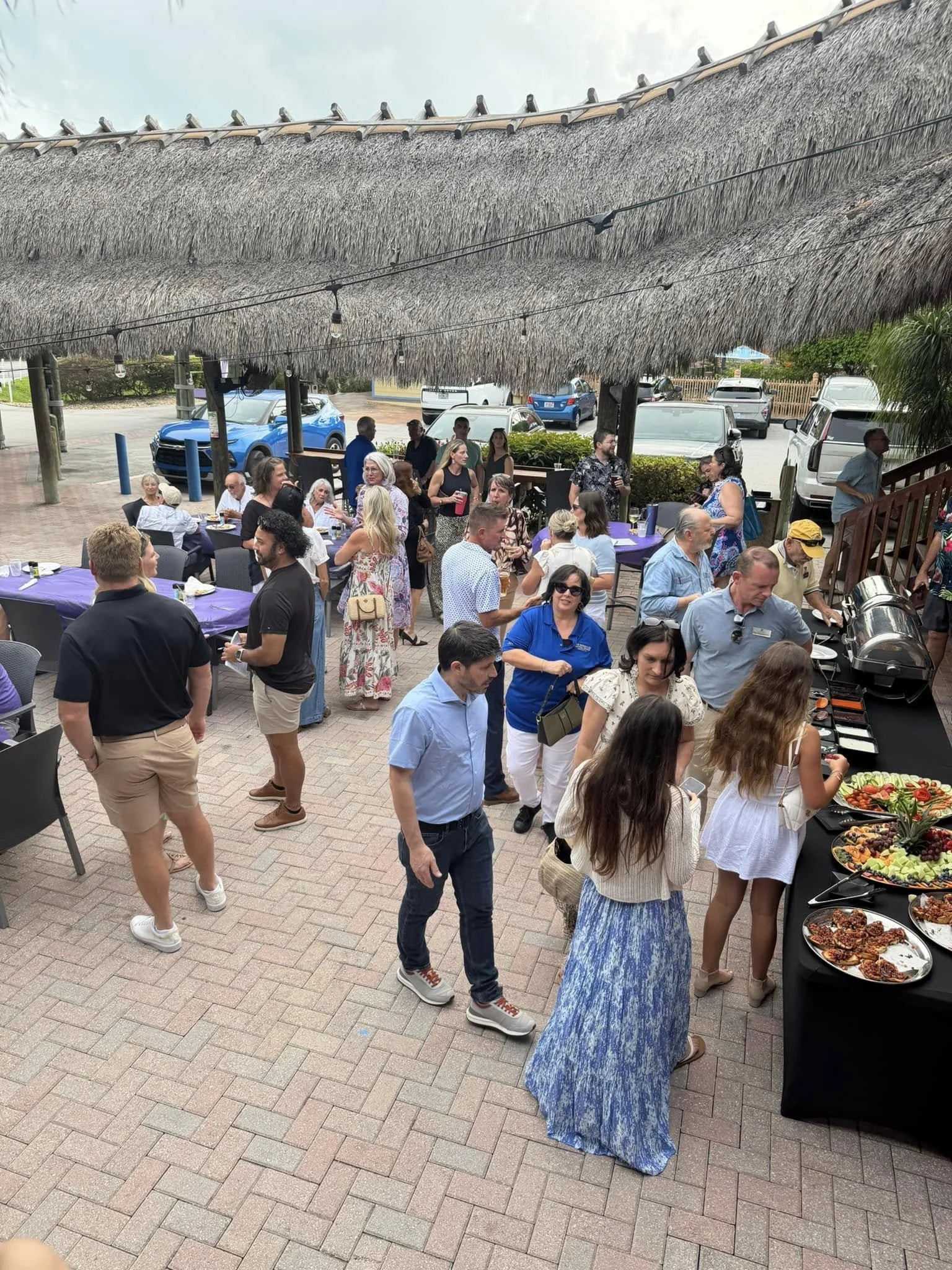 People at an outdoor buffet with food on a table, some in line, and others mingling under a thatched roof. Parking lot with cars is visible in the background.