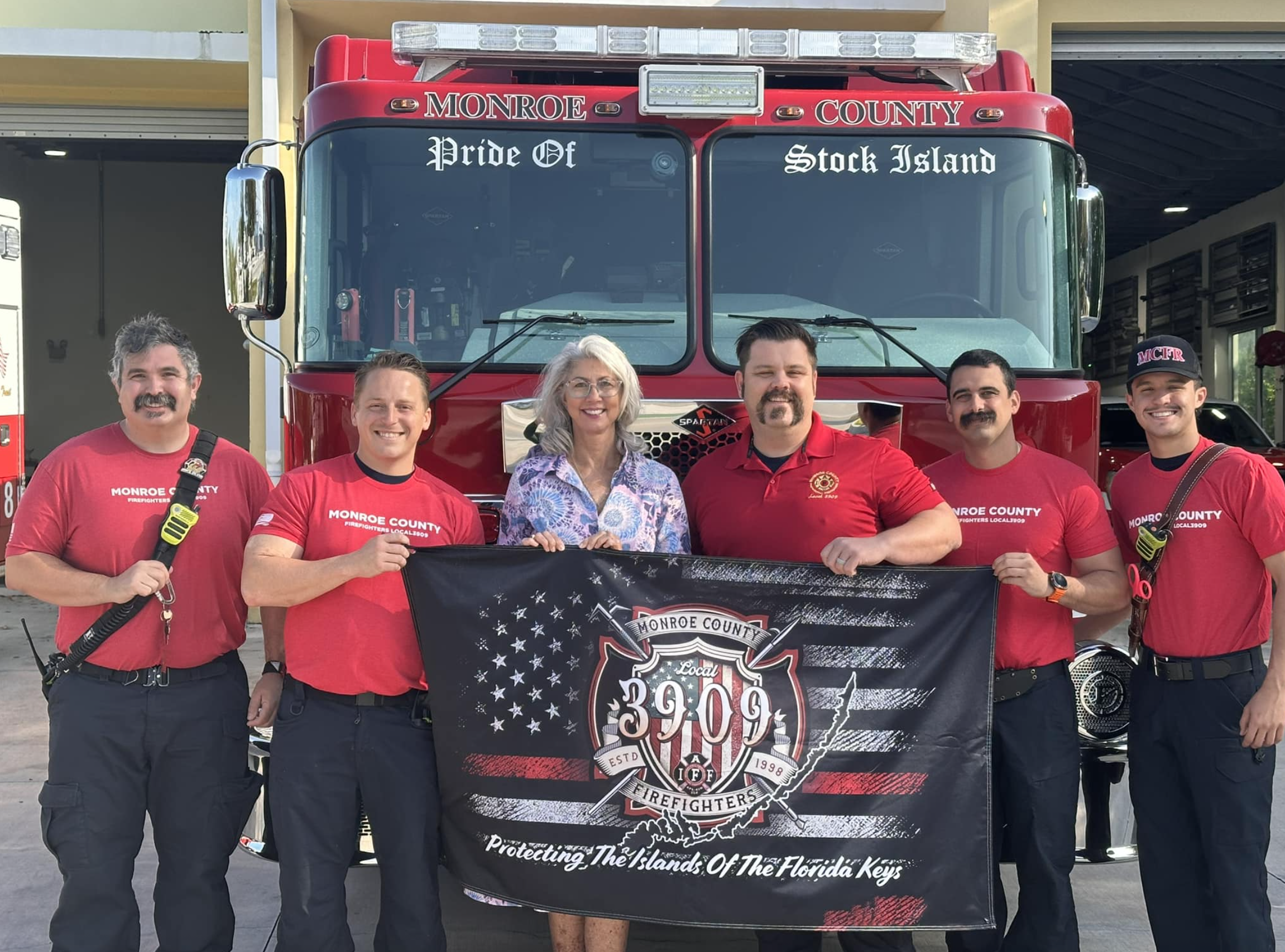 Group of six firefighters and a woman holding a flag in front of a red fire truck labeled Monroe County, Pride Of Stock Island, Florida Keys. Michelle Lincoln