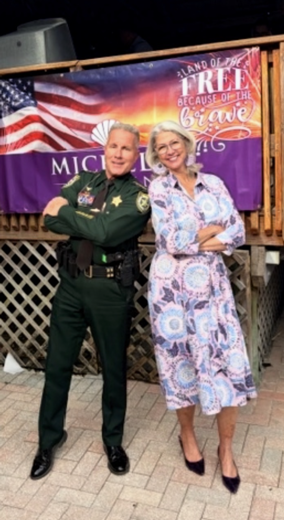 A man dressed as a sheriff in a green uniform and a woman in a patterned dress standing side by side with arms crossed, smiling, in front of a banner with an American flag, sunset, and the words 'Land of the Free Because of the Brave'.