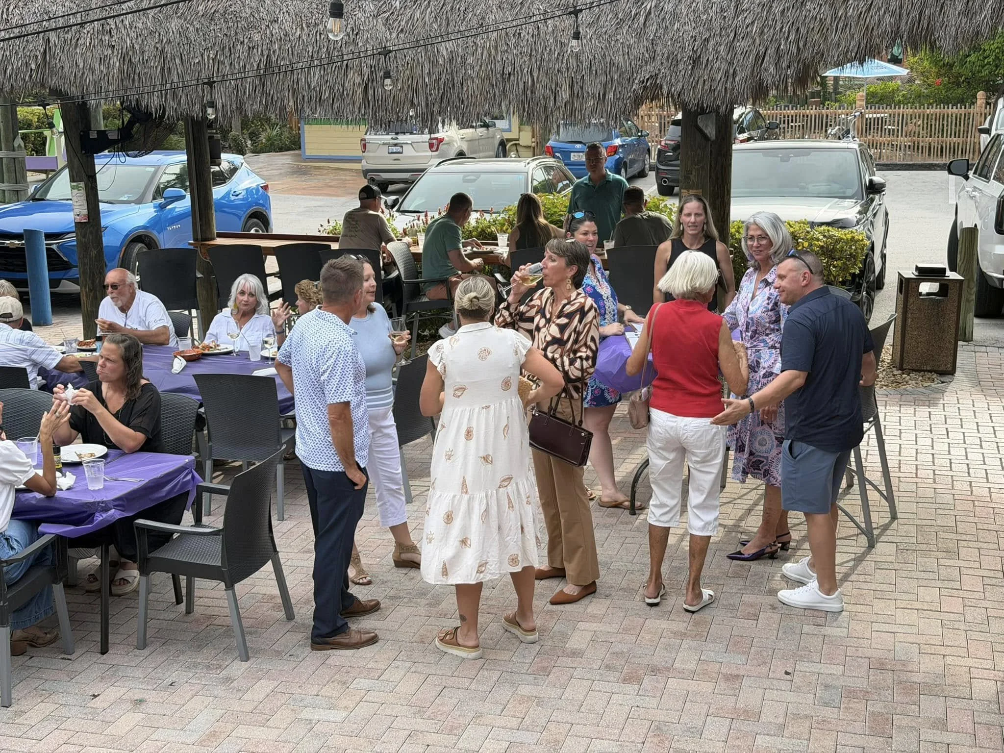 Group of people socializing outdoors under a thatched roof at a patio, some sitting at tables with purple tablecloths, others standing and chatting, with parked cars and a fence visible in the background.