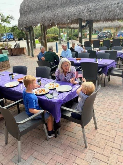 People dining outdoors at a restaurant with purple tablecloths, under a thatched roof structure, enjoying food and drinks, with a woman holding an American flag and children sitting at the table.