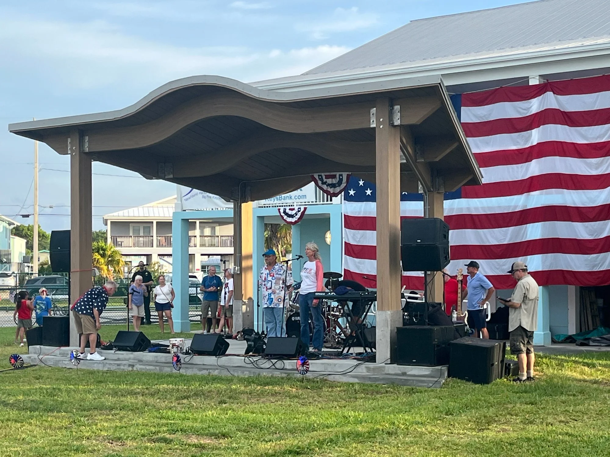 People setting up on a stage with a large American flag backdrop, in an outdoor park on a sunny day.