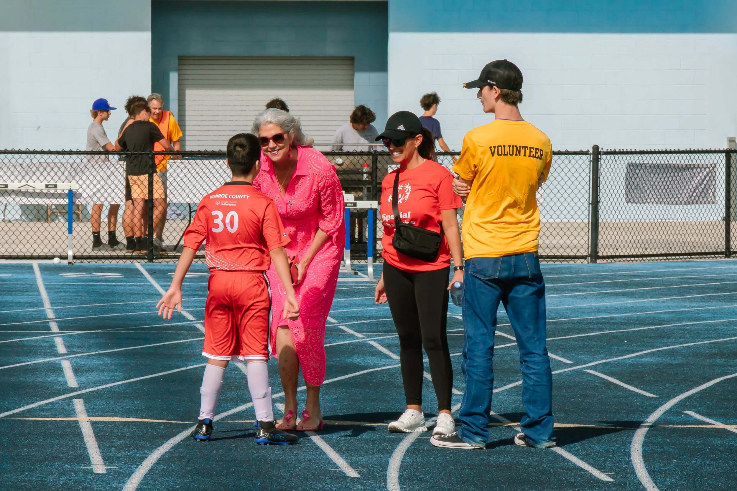 A group of people on a running track at an outdoor sports event, including a young boy in a red sports uniform, two women wearing red shirts and black pants, one with a black cap and the other with sunglasses, and a volunteer in a yellow shirt and blue jeans. The older woman is smiling at the boy.