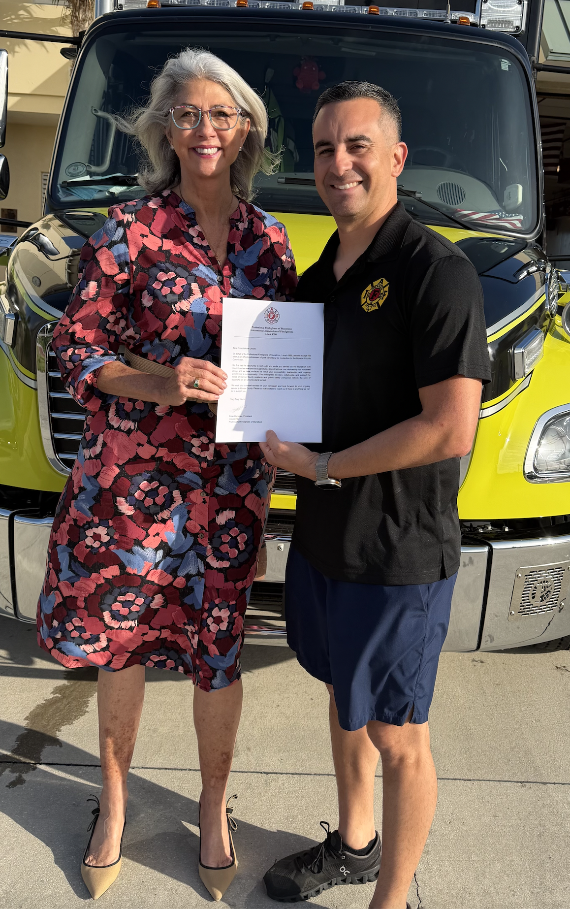 A woman with gray hair and glasses holding a document while standing next to a man in a black shirt and shorts in front of a fire truck. They are smiling.