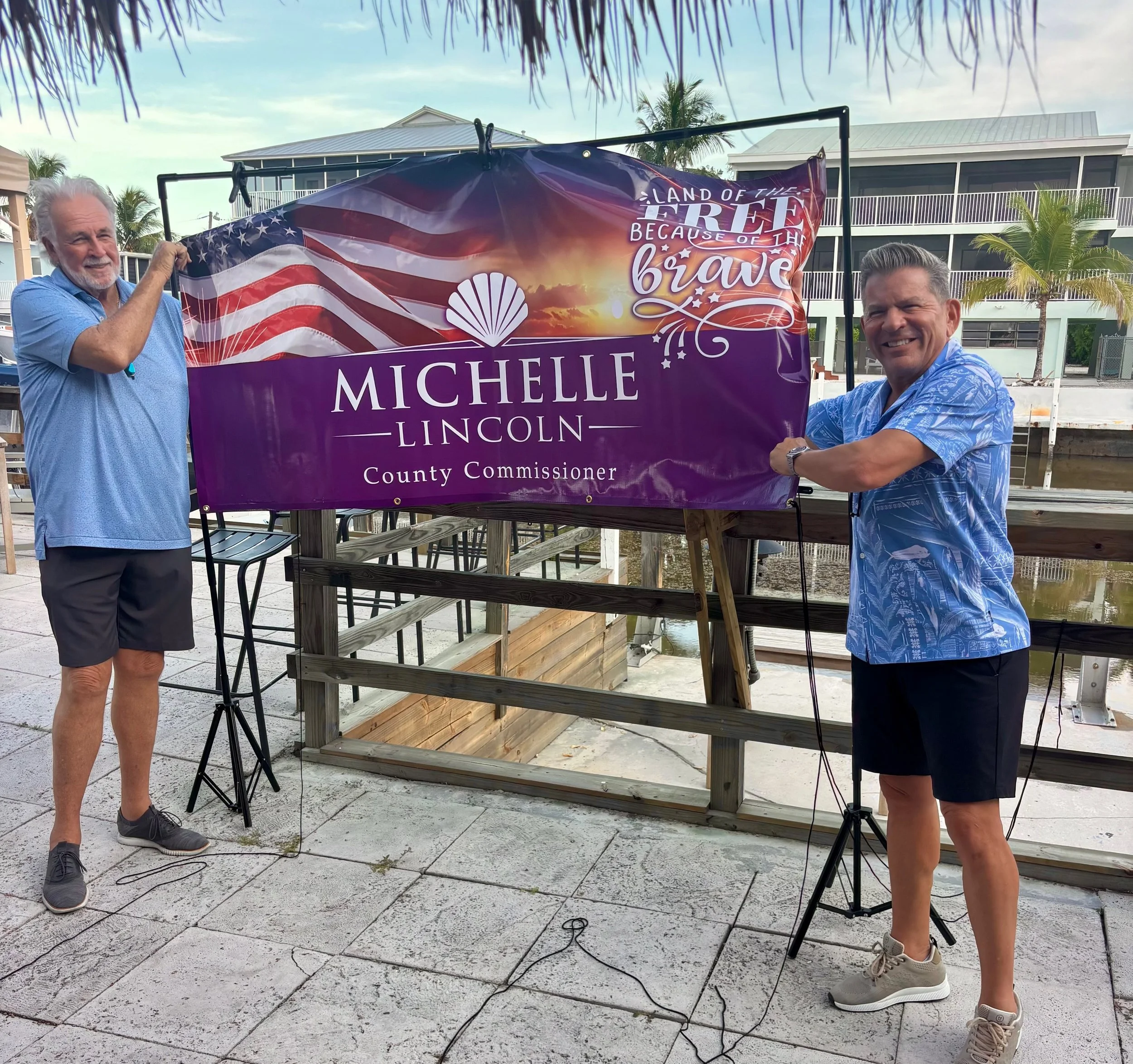 Two men holding a purple sign with an American flag design and the text 'Land of the free because of the brave, Michelle Lincoln, County Commissioner' at an outdoor venue with palm trees and residential buildings in the background.