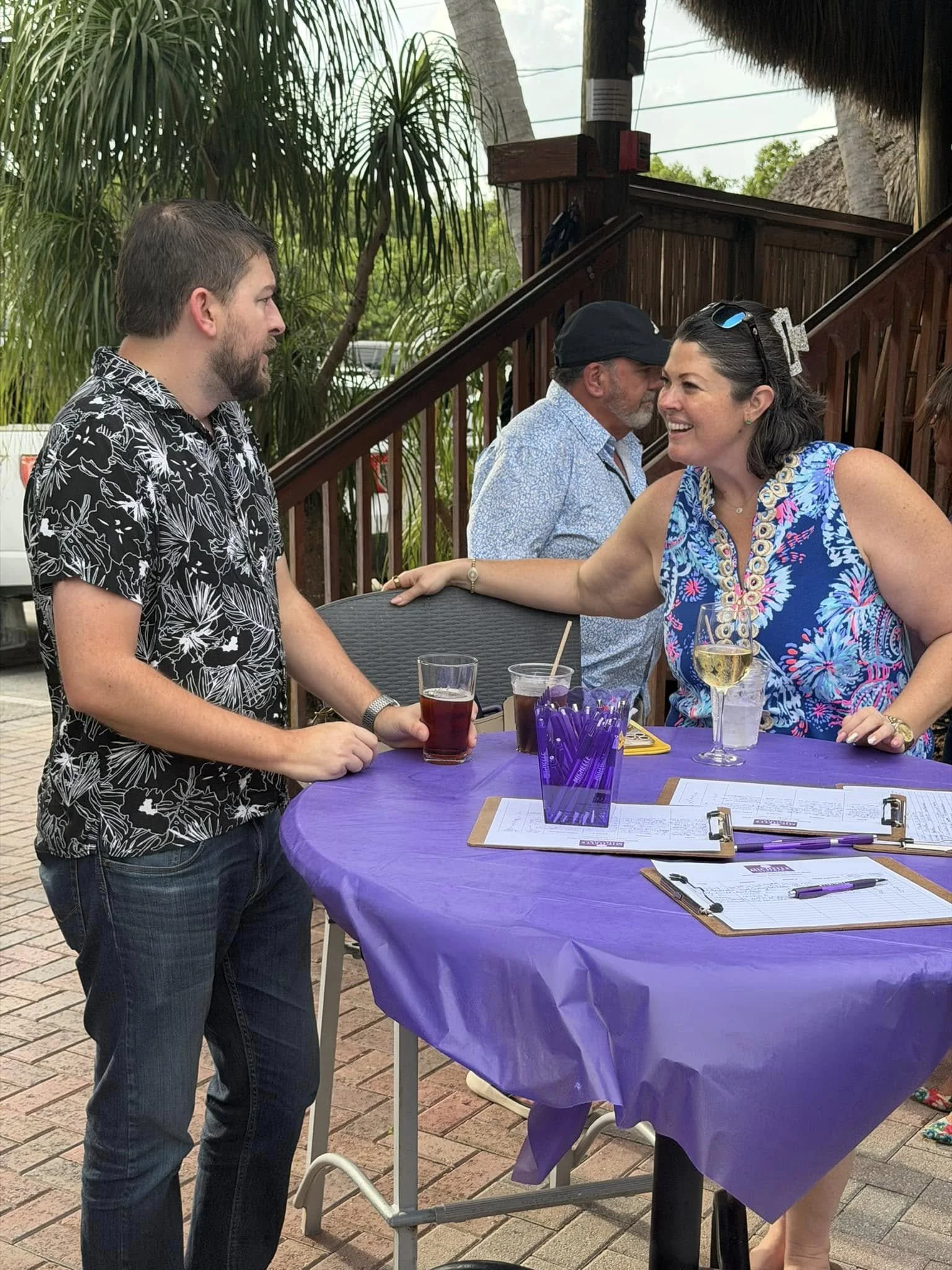 A woman and a man are standing near a purple table outdoors, smiling and engaging in conversation. The woman has dark hair, is wearing a colorful sleeveless dress, and has a glass of wine in front of her. The man has a beard, short hair, and is weari