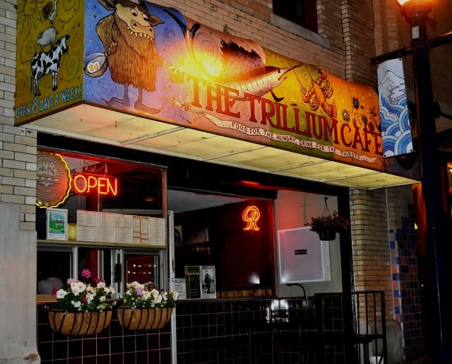 The Trillium Cafe storefront with a colorful cartoon-style sign, neon 'OPEN' sign, and flower baskets outside.