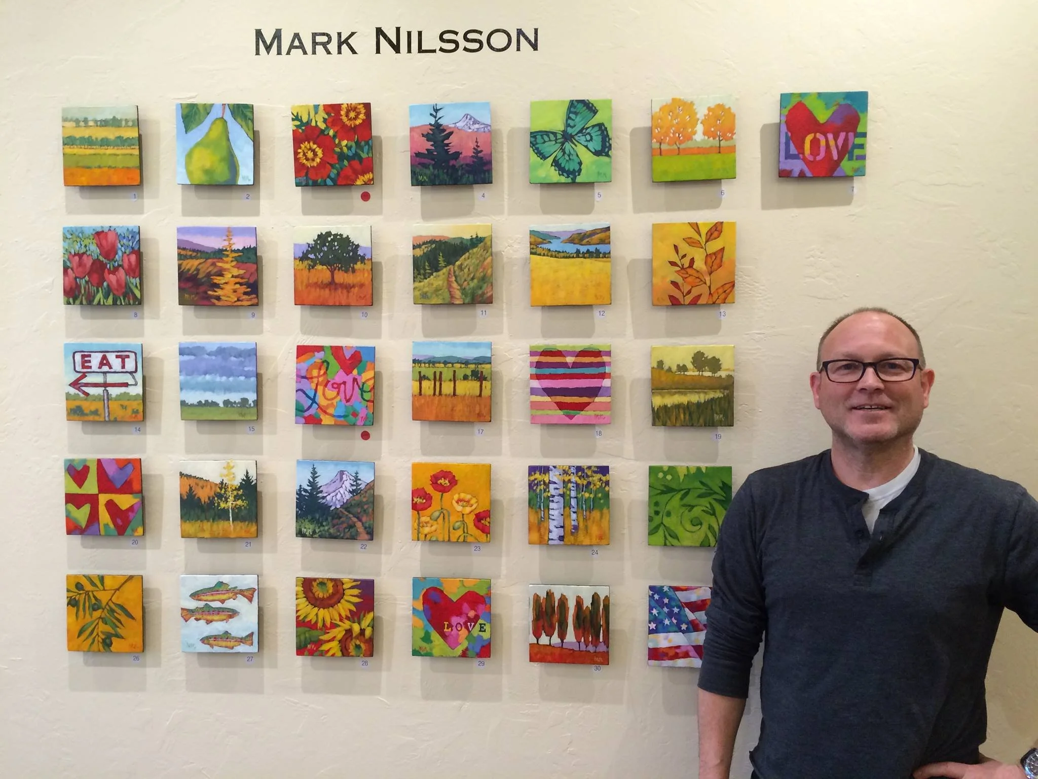 A man smiling and wearing glasses stands to the right of a wall display with 30 small, colorful paintings arranged in a grid, titled 'Mark Nilsson'.