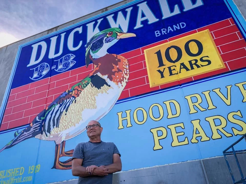 A man in glasses and a gray t-shirt standing in front of a large mural advertising Duckwalters, a brand celebrating its 100th anniversary, featuring a colorful duck and the text 'Duckwalters 1919-2019, 100 Years, Hood River Pears'. Mark Nilsson ©