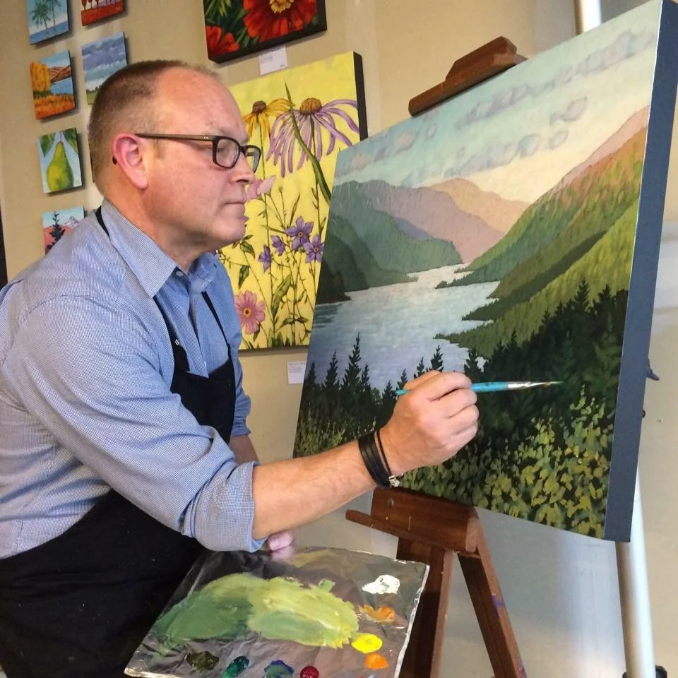 A Mark Nilsson is painting a landscape scene of mountains, river, and trees on a canvas at an art gallery. Mark Nilsson ©