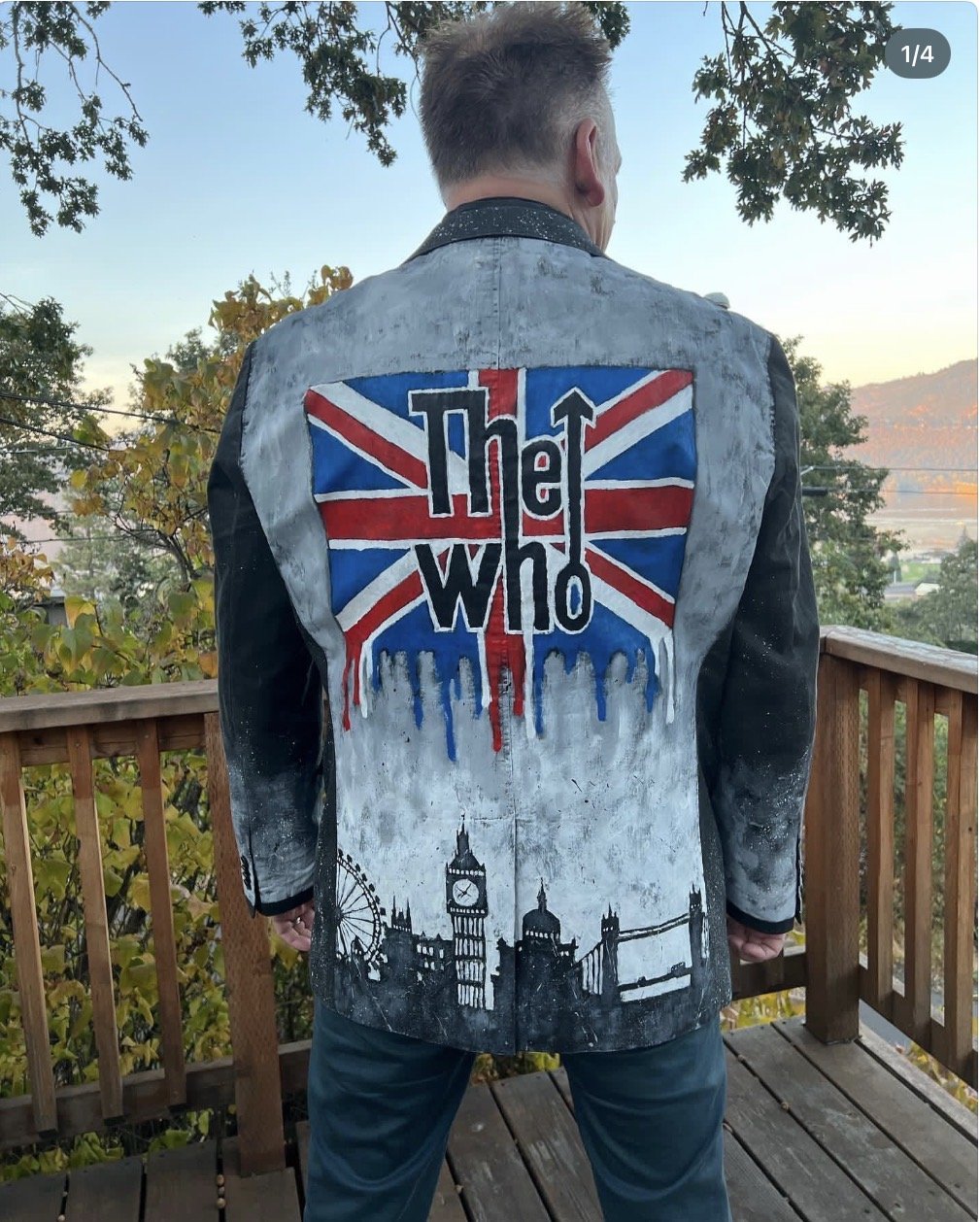 A person standing outdoors on a wooden deck, facing away, wearing a jacket with a painted British flag, the words 'The Who,' and silhouettes of London's landmarks like Big Ben and Tower Bridge, Mark Nilsson ©