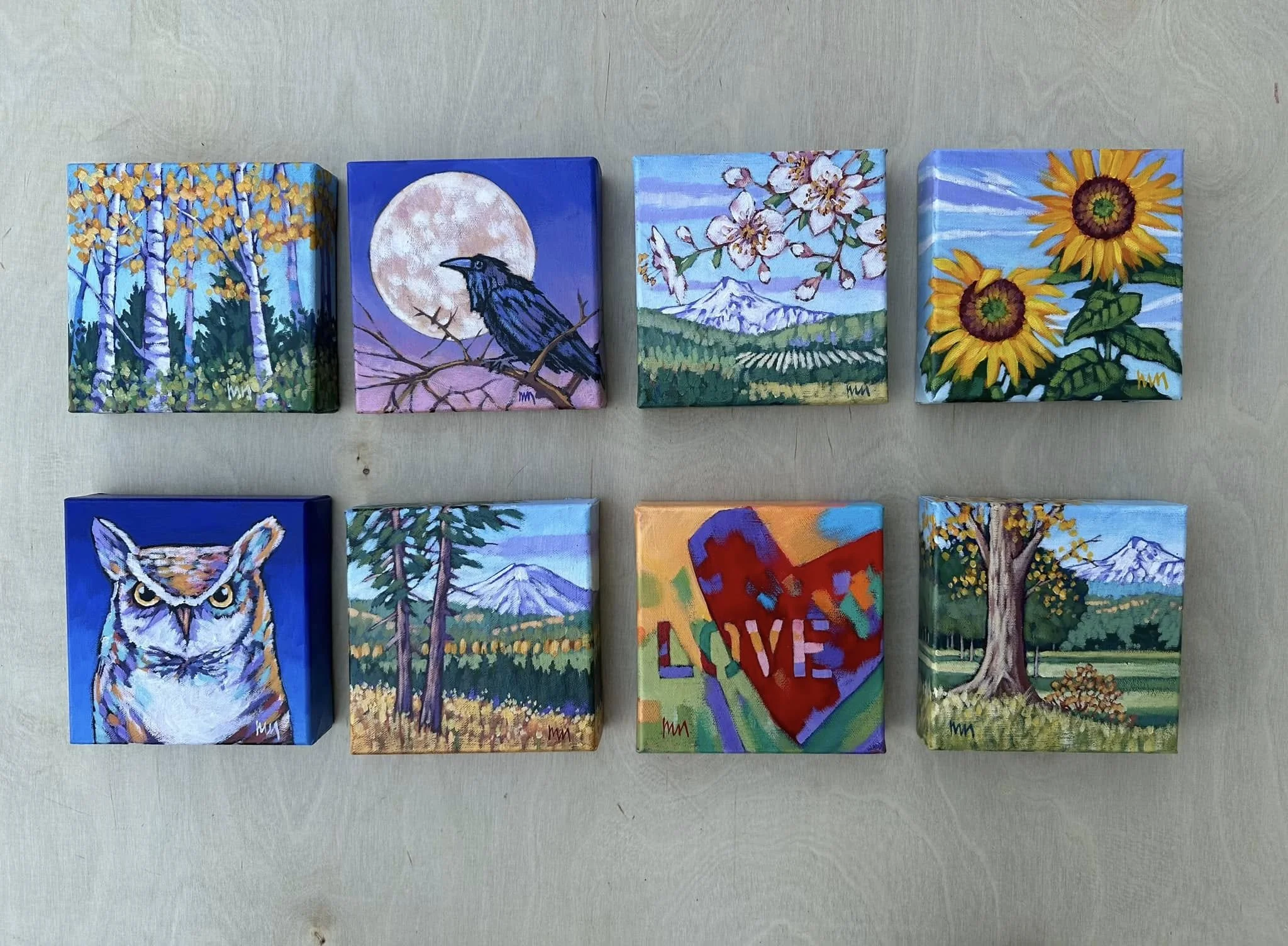 A collection of eight small painted canvases arranged on a light wooden surface, each depicting various scenes including trees, animals, flowers, mountains, a moon, and a colorful abstract piece with the word 'LOVE'. Mark Nilsson ©