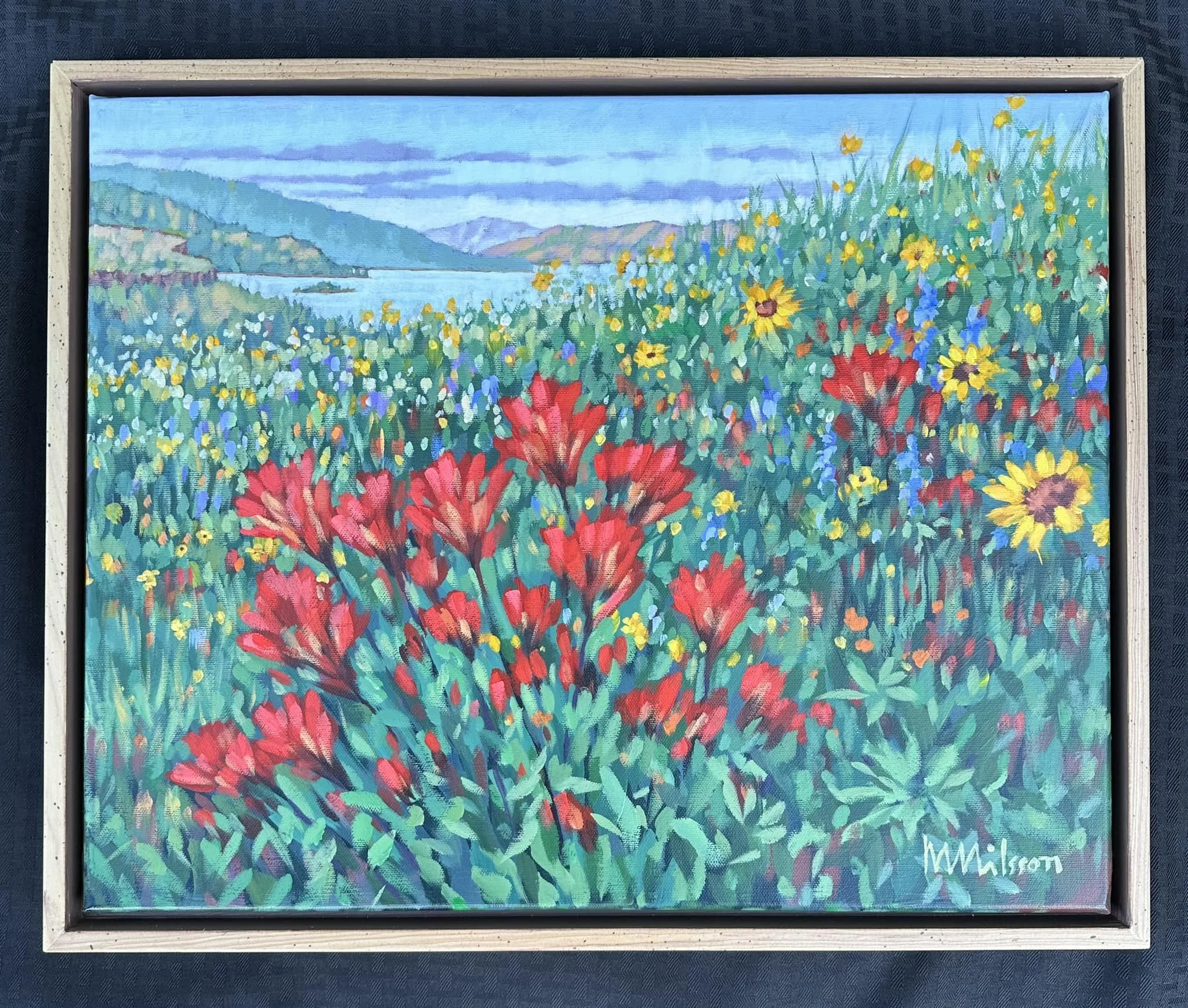 A colorful landscape painting of a lakeside scene with a field of red, yellow, and blue flowers in the foreground, a calm lake and distant mountains in the background, under a cloudy sky. Mark Nilsson ©