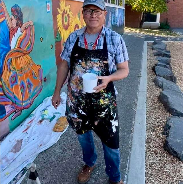 standing outdoors in front of a colorful mural, wearing an apron splattered with paint, holding a white container, and smiling at the camera.  Mark Nilsson ©