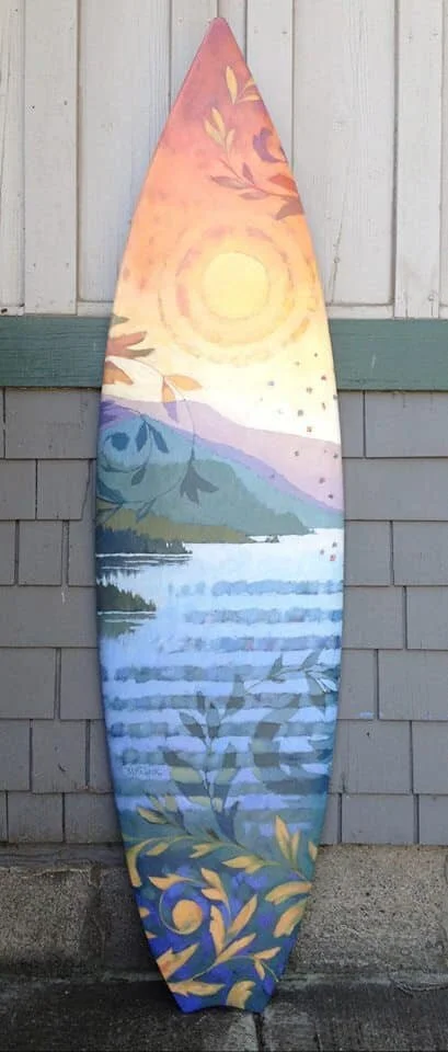 Colorful painted surfboard with a landscape scene of a sunset over mountains, water, and foliage, leaning against a white wooden wall with a gray base. Mark Nilsson ©